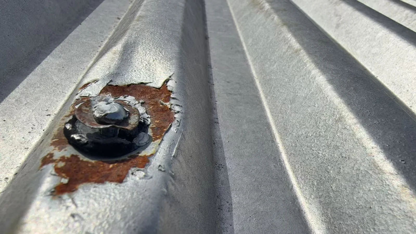 Close view of a corroded screw and worn washer on an aged galvanised metal roof rib